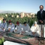 Hombres de blanco sentados en alfombras en un acto al aire libre con la Alhambra al fondo