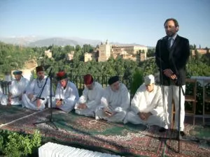 Hombres de blanco sentados en alfombras en un acto al aire libre con la Alhambra al fondo