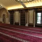 Sala de oración con alfombra granate en hileras, puertas de madera y el mihrab al fondo