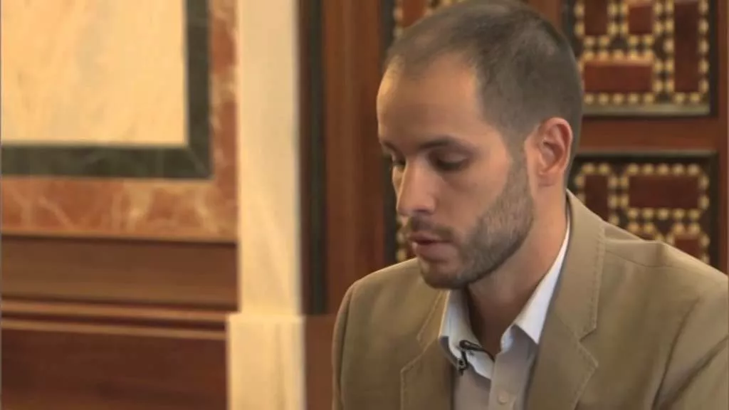 Hombre joven con chaqueta beige y micrófono de solapa habla sentado en el interior de la mezquita