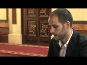 Hombre de chaqueta oscura hablando sentado ante las puertas de madera taraceada y la alfombra roja