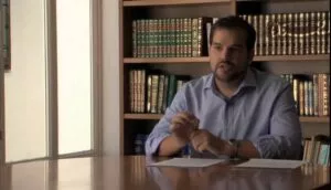Hombre hablando sentado a una mesa ante una estantería de libros islámicos