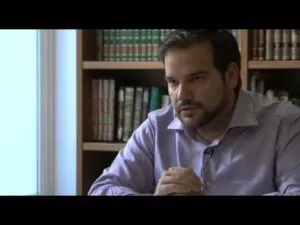 Hombre con camisa lila sentado ante una estantería de libros, hablando con las manos juntas