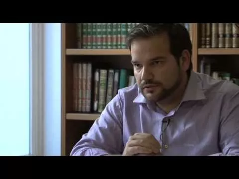Hombre con camisa lila sentado ante una estantería de libros, hablando con las manos juntas