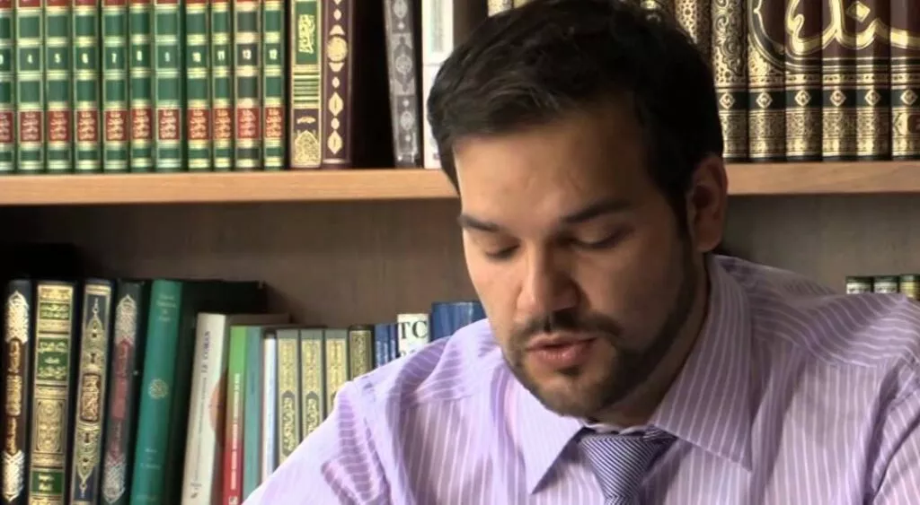 Hombre con camisa de rayas y corbata lee unos papeles ante una estantería de libros
