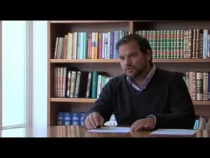Plano general de un hombre sentado a una mesa hablando, con estanterías de libros detrás