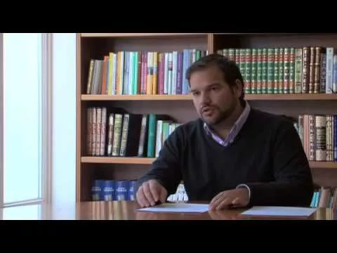 Plano general de un hombre sentado a una mesa hablando, con estanterías de libros detrás