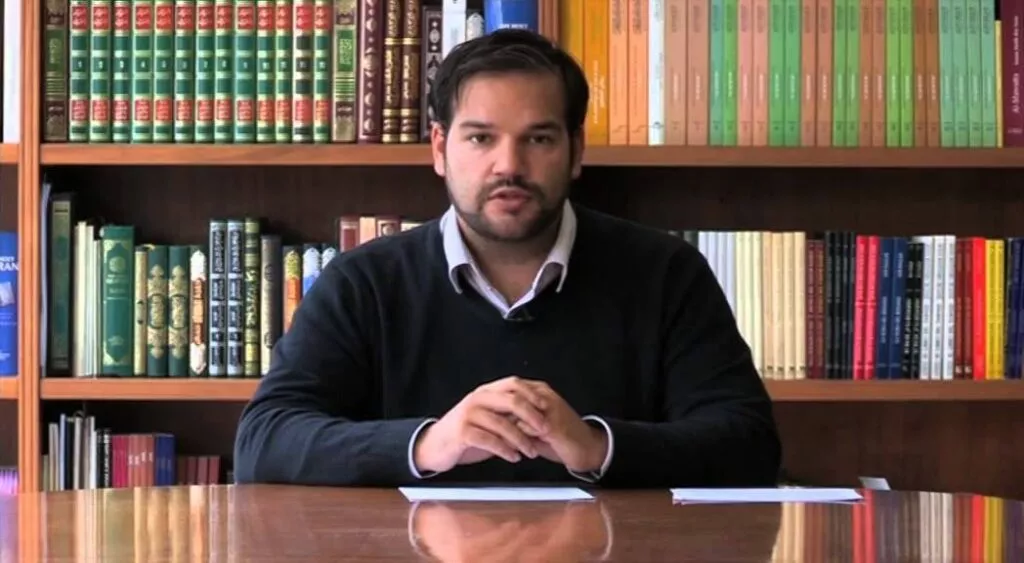 Un hombre joven habla a cámara sentado a una mesa, ante una estantería llena de libros
