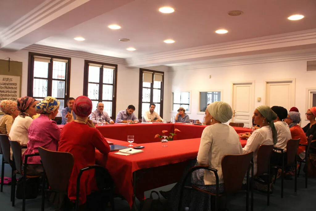 Mujeres con pañuelo y ponentes en torno a mesas con mantel rojo durante un seminario