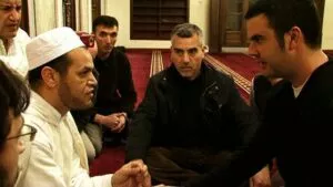 Hombre con gorro blanco estrecha la mano de un joven en la sala de oración durante la shahada