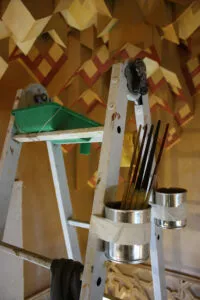 Escalera de pintor con pinceles y botes atados, ante el techo de mocárabes de madera pintada del zaguán