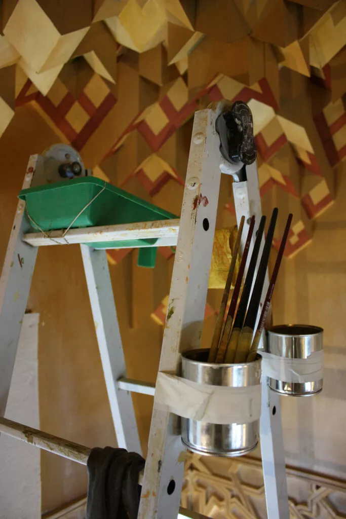 Escalera de pintor con pinceles y botes atados, ante el techo de mocárabes de madera pintada del zaguán