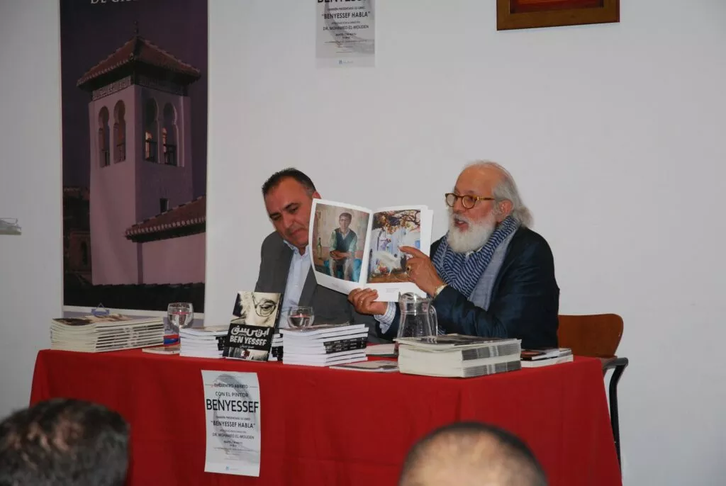 El pintor Benyessef muestra las láminas de su libro junto al presentador en la mesa del encuentro