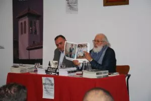 El pintor Benyessef muestra las láminas de su libro junto al presentador en la mesa del encuentro