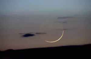 Fina luna creciente al atardecer sobre un cielo azulado con nubes bajas