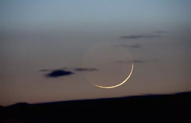 Fina luna creciente al atardecer sobre un cielo azulado con nubes bajas