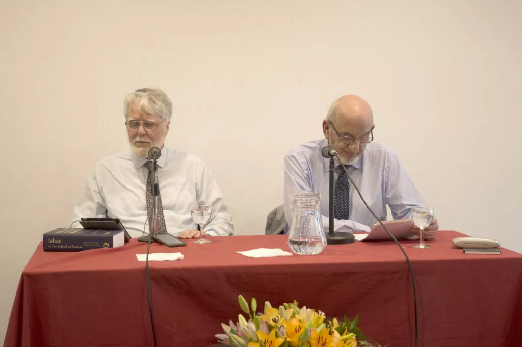 Dos ponentes ante los micrófonos en una mesa con mantel rojo y un libro sobre el Islam