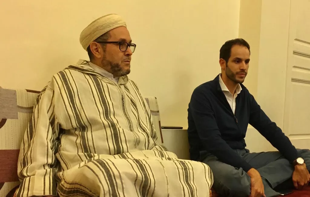 Ulema con turbante y chilaba a rayas sentado en un sofá junto a un joven durante una visita
