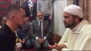 Imam con turbante blanco habla con un joven sentado frente a él, rodeados de asistentes en la mezquita