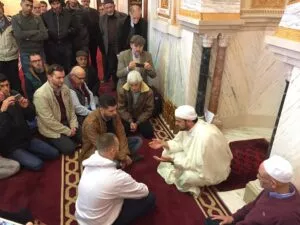 Imam de blanco sentado en la alfombra roja habla ante un corro de hombres junto a las columnas de mármol