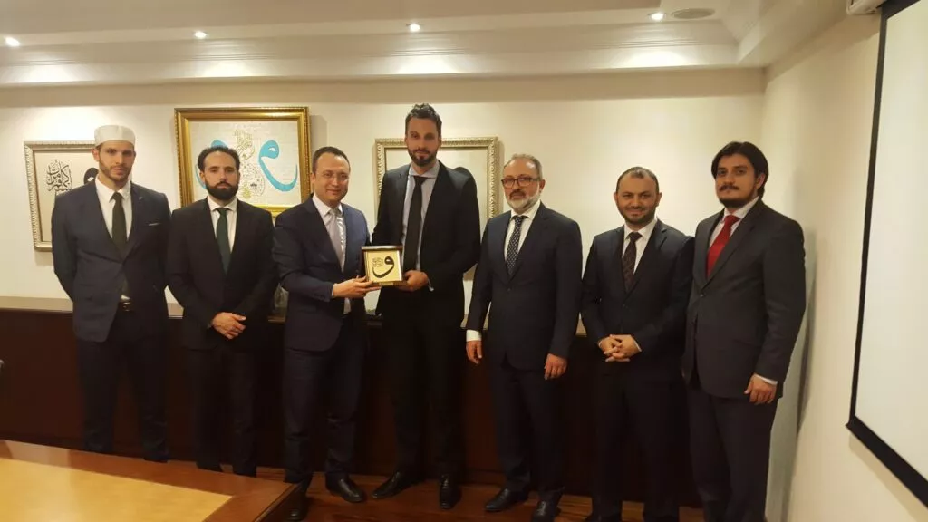 Siete hombres trajeados posan en la entrega de una placa dorada con caligrafía árabe