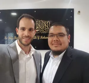 Dos hombres posan ante un panel negro con la shahada en caligrafía dorada y su traducción al español