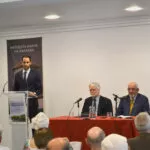 Un joven presenta en el atril a dos ponentes sentados a la mesa bajo el rótulo Mezquita Mayor de Granada