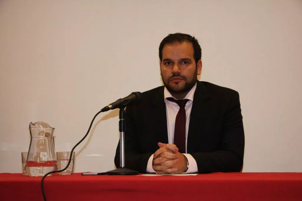 Hombre con traje oscuro y corbata granate sentado tras una mesa con micrófono y jarra de agua
