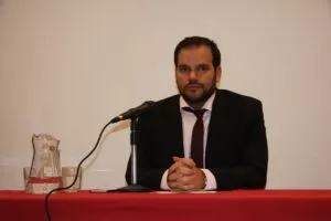 Hombre con traje oscuro y corbata granate sentado tras una mesa con micrófono y jarra de agua