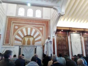 Predicador de blanco lee la jutba ante el mihrab mientras los fieles escuchan sentados