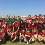 Grupo de adolescentes con camisetas granates de un campamento posando en el césped junto a una ría