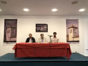 Tres ponentes en una mesa con mantel rojo entre las banderolas de la Mezquita Mayor de Granada