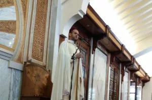 Imam con chilaba blanca y gorro rojo predica ante el micrófono junto al mihrab de madera tallada