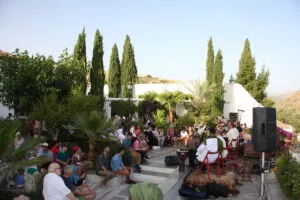 Público sentado en un jardín con cipreses escucha a músicos vestidos de blanco con tambores