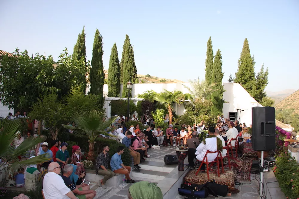 Público sentado en un jardín con cipreses escucha a músicos vestidos de blanco con tambores