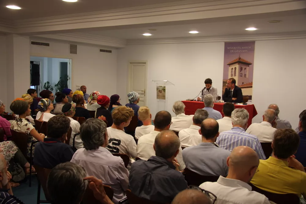 Sala llena durante una ponencia, con los dos ponentes en la mesa y un atril junto al cartel de la Mezquita Mayor