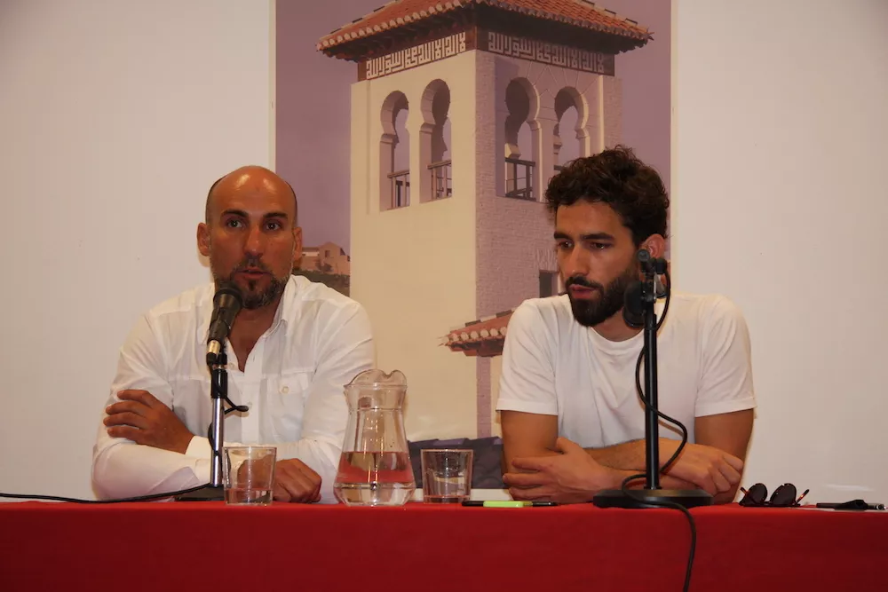 Dos ponentes hablan ante los micrófonos en la mesa, con el cartel del alminar de la mezquita al fondo
