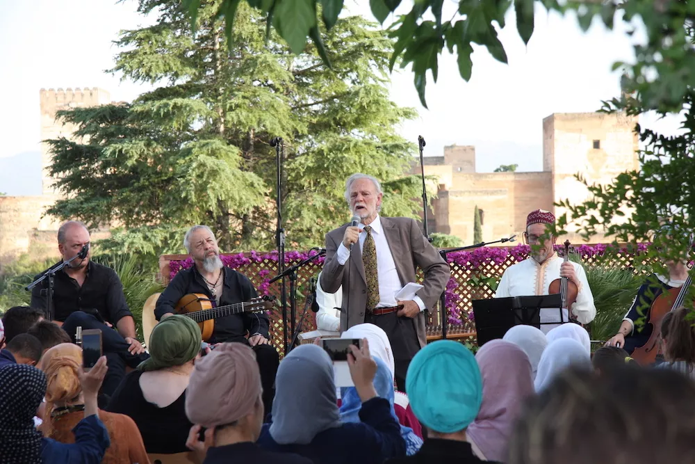 Concierto en el jardín: un presentador habla al micrófono junto a los músicos con guitarra y violín, con la Alhambra al fondo