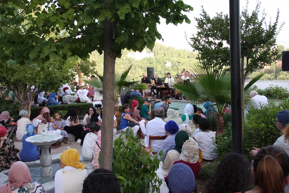 Público sentado en el jardín escuchando al grupo de música, con la sierra al fondo