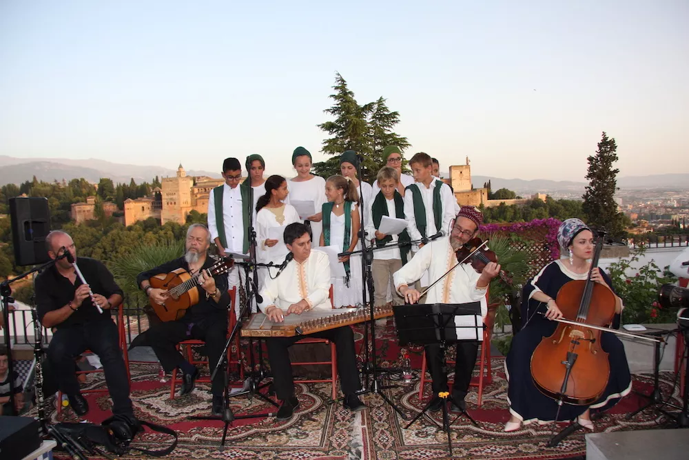 Grupo musical con laúd, cítara, violín, chelo y flauta acompañado de un coro de niños, con la Alhambra al fondo