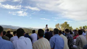 Fieles sentados al aire libre en el monte escuchan la jutba de un hombre vestido de blanco