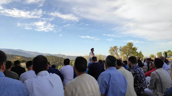 Fieles sentados al aire libre en el monte escuchan la jutba de un hombre vestido de blanco