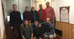Ocho hombres posan en una sala pequeña junto a un cartel de invocaciones en árabe