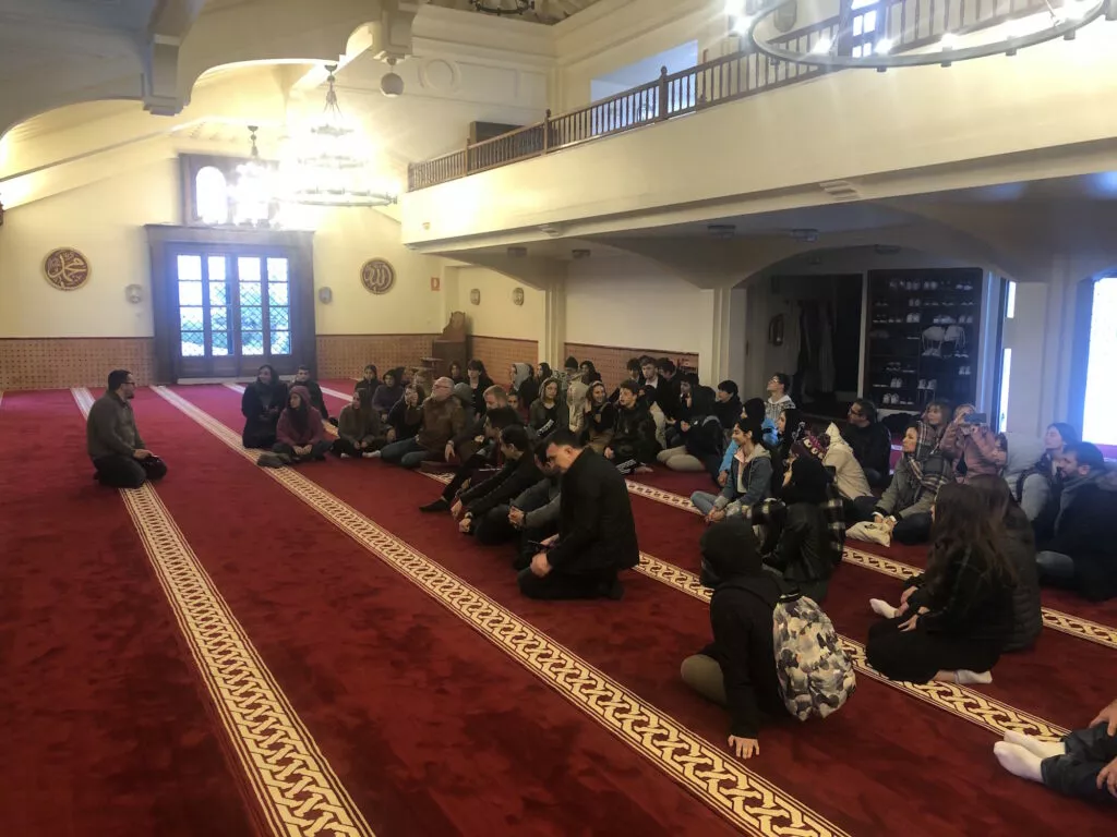 Estudiantes sentados en la alfombra roja de la sala de oración escuchando la explicación