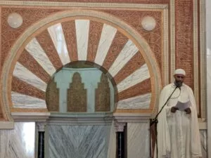 Predicador de blanco leyendo la jutba junto al gran arco de herradura rojo y blanco del mihrab