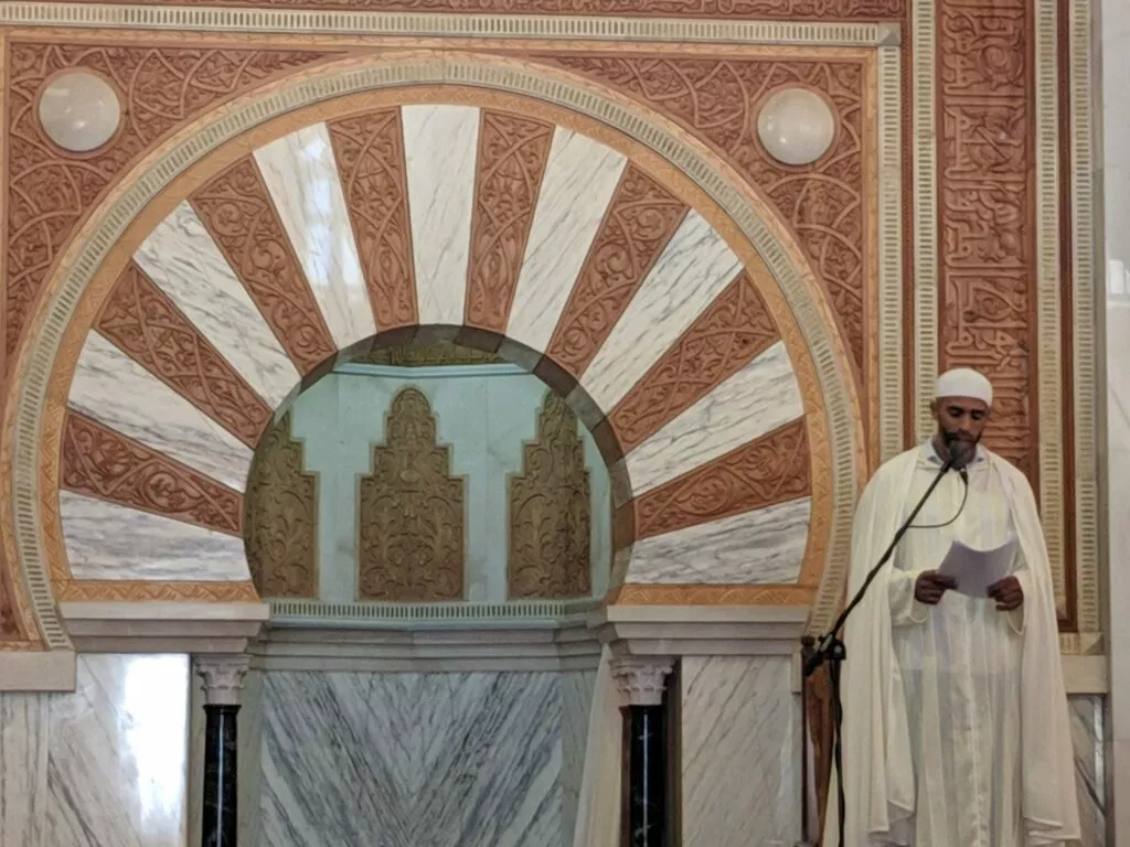 A preacher dressed in white reads the khutba before the mihrab of red arches and white marble