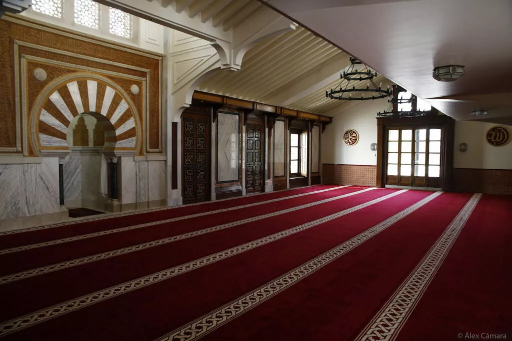 Sala de oración vacía con el mihrab dorado a la izquierda y las alfombras rojas en hileras