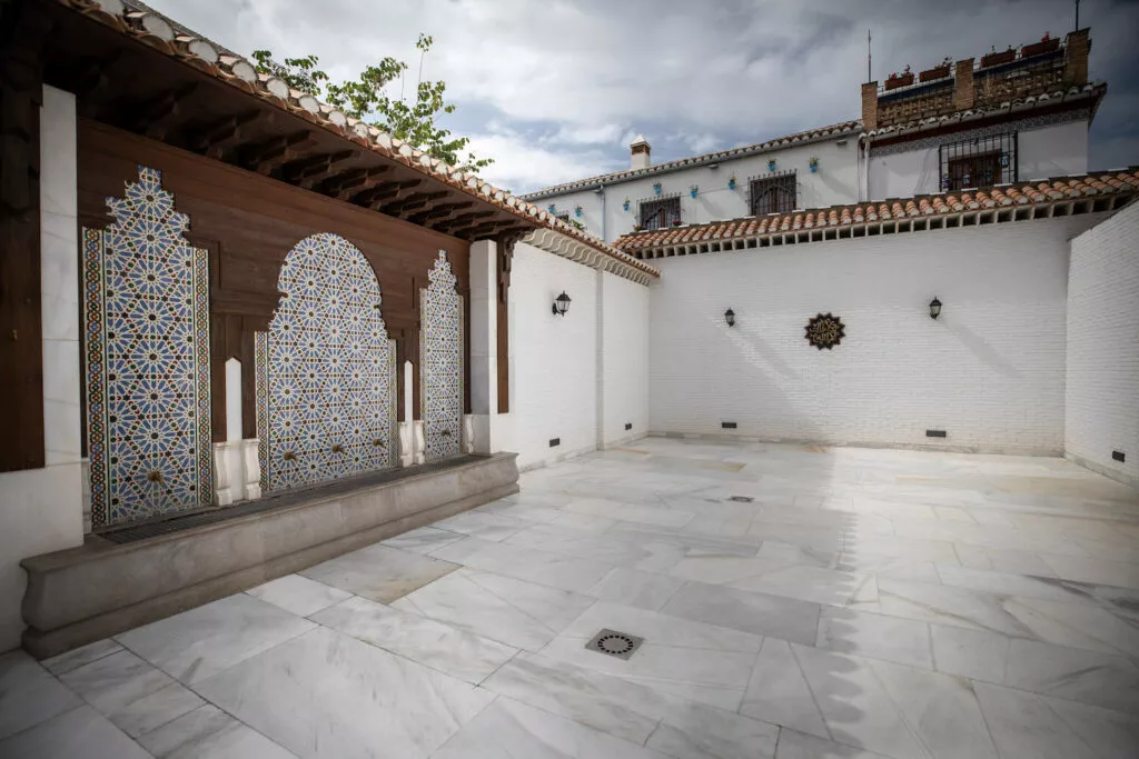 Patio de abluciones con la fachada de madera y los paneles de alicatado, y el Albaicín asomando sobre la tapia