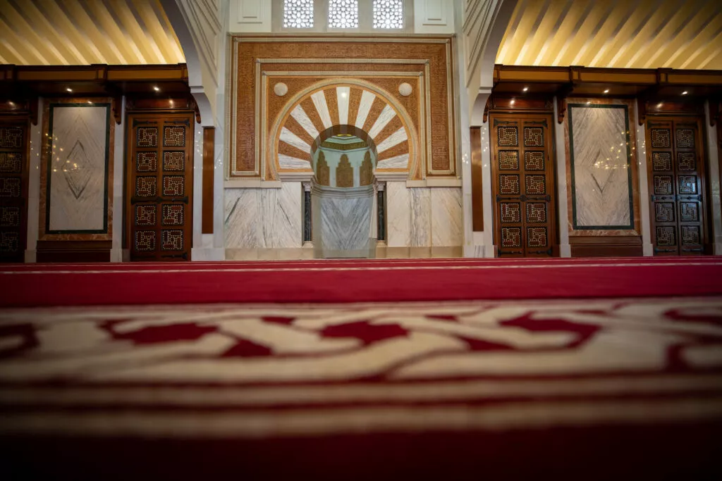 Sala de oración a ras de la alfombra roja, con el mihrab al fondo entre puertas de madera