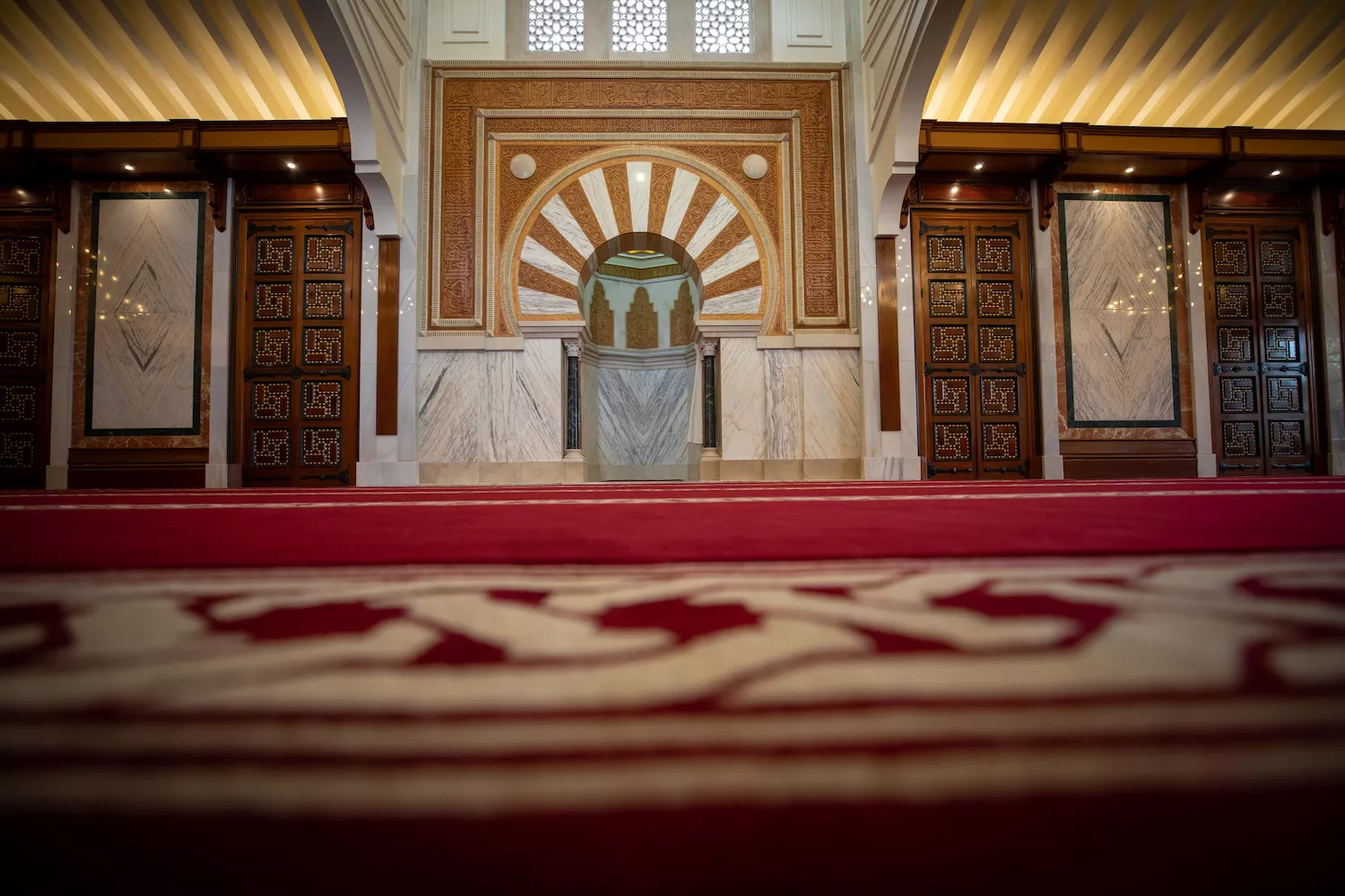 Sala de oración a ras de la alfombra roja, con el mihrab al fondo entre puertas de madera